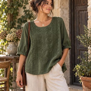 Puede incluir: Una blusa verde oliva con cuello redondo, mangas tres cuartos y un panel central con intrincados bordados florales y detalles de encaje, combinada con pantalones de color beige claro y un bolso de hombro de cuero marrón.