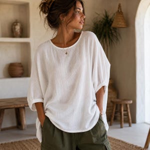 May include: A loose-fitting white blouse with a round neckline and three-quarter length sleeves, featuring a textured, crinkled fabric finish. The garment is paired with olive green trousers that have visible pockets, creating a relaxed silhouette.