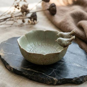 May include: A light green ceramic bowl with a scalloped edge and a textured spiral pattern on the interior base. Two small, sculpted ceramic birds are attached to the rim of the bowl, facing inward. The bowl has a speckled, matte finish.