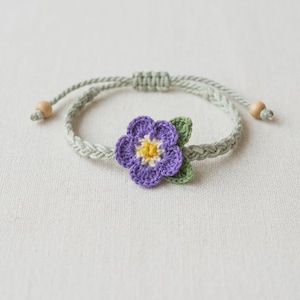 Peut inclure: Un bracelet crocheté à la main avec une fleur violette au centre jaune et des feuilles vertes. Le bracelet a une bande tressée vert clair, des perles en bois et une fermeture réglable.