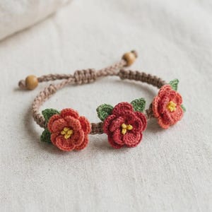 May include: A handmade crochet bracelet featuring three red and orange flower appliqués with green leaves and yellow centers. The bracelet is adjustable with a tan cord and wooden beads, perfect for a floral accessory.