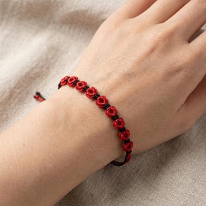 May include: A bracelet featuring a series of small, red, flower-shaped knots linked together by a thin black cord. The design consists of repeating red floral motifs separated by black knots, with adjustable black and red cord ends visible at the clasp.