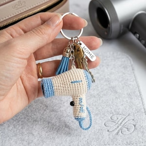 May include: A cream and light blue crochet hairdryer keyring. The keyring includes a blue tassel, keys, and a small "HOME" tag. The miniature hairdryer has the word "on/off" on it. The keyring is attached to a silver ring.