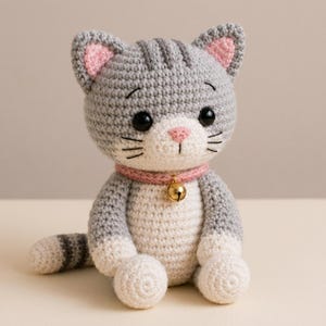 Puede incluir: Una figura de gato tejida a ganchillo con hilo gris y blanco, con orejas rosadas por dentro, un collar rosa y un pequeño cascabel dorado. El gato tiene bigotes negros bordados, una nariz rosa y grandes ojos negros de plástico.