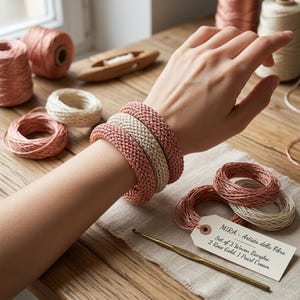 May include: Three woven bangles in rose gold and pearl cream colors are displayed on a wrist. The image also shows spools of thread, a crochet hook, and a tag that reads "MIRA - Artista della Fibra".