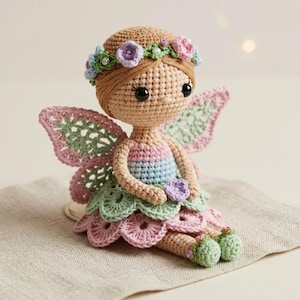May include: A handmade crochet fairy doll with a floral crown and delicate wings. The doll features a pastel color palette of pink, green, and blue. The fairy is seated on a neutral-colored surface.