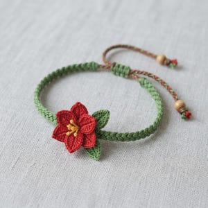 May include: A handmade crochet bracelet featuring a vibrant red flower with yellow center and green leaves. The bracelet has a green braided band and adjustable closure with wooden beads. A unique, floral accessory.