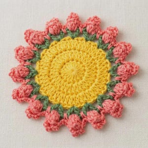 May include: A handmade crochet coaster in the shape of a flower. The center is yellow, surrounded by pink petals and green leaves. The coaster is made of yarn and is likely used for placing cups or mugs.