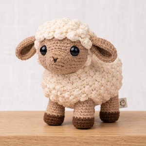 May include: A handmade crochet sheep toy with a cream-colored body, brown face, ears, and hooves. The sheep has large black eyes and is standing on a wooden surface. A small tag with a sheep icon is attached.