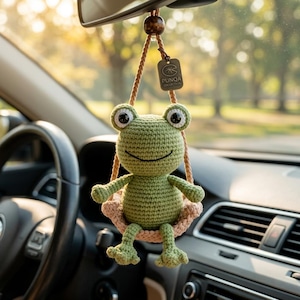 Op de afbeelding: Een groene gehaakte kikker auto-hanger met grote ogen en een lachende mond, hangend aan een bruin touwtje. De kikker zit in een lichtroze gehaakte schommel. Een klein label met "PUNQA COLLECTION" is zichtbaar.
