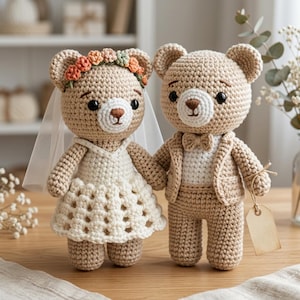 May include: Two crocheted teddy bears, a bride and groom, stand side-by-side. The bride bear wears a white dress, veil, and floral crown. The groom bear is in a suit with a bow tie. They are holding hands, perfect for a wedding or anniversary gift.