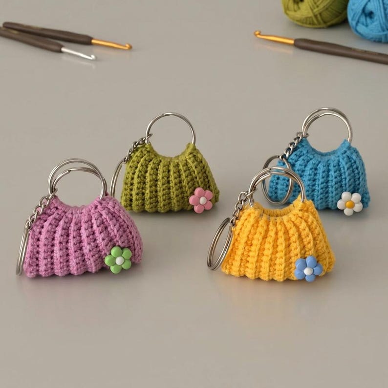 May include: Four colorful crochet purse keychains. The keychains are in shades of pink, green, yellow, and blue, each with a silver keyring and a small flower accent. Crochet hooks and yarn are visible in the background.