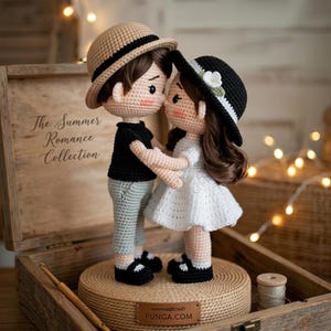May include: Crocheted dolls of a couple embracing, wearing hats and standing on a woven platform. The male doll wears a black shirt and grey trousers, while the female doll wears a white dress. The dolls are part of "The Summer Romance Collection."