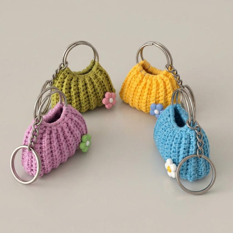 May include: Four handmade crochet purse keychains in green, yellow, purple, and blue. Each mini purse has a silver-tone ring and chain, and a small flower accent. The keychains are displayed on a light background.