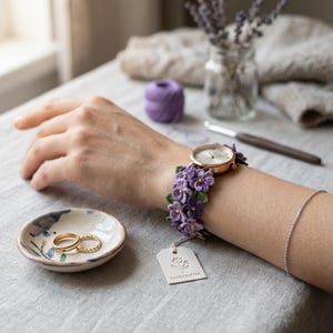 May include: A gold-tone watch with a white face and a band adorned with purple crochet flowers. A small ceramic dish with two gold rings sits nearby. A tag that says "HANDCRAFTED" hangs from the watch.