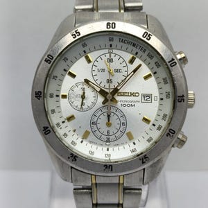 May include: A silver and gold-tone Seiko chronograph watch with a white dial, three sub-dials, and a tachymeter bezel. The watch face displays the text Seiko, Chronograph, 100M, and a date window showing the number 12. It has a metal link band.