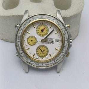 May include: A silver Seiko Quartz Sports 150 watch with a white face, gold detailing, and several subdials. The watch has a date display and a bezel marked with city names. The watch is presented on a neutral background.