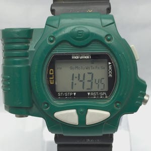 May include: A green digital watch with a black band. The watch face displays the time as 1:43:45 and the day of the week. The brand name "maruman" is visible above the digital display. The watch has white buttons and a black strap.