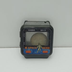 Può includere: Orologio digitale vintage Casio GH-17 Space Hero. L'orologio ha una cassa nera con un quadrante blu e arancione. Il quadrante presenta le parole "SPACE HERO" e una grafica di un astronauta. Lo schermo mostra un oggetto di colore chiaro.