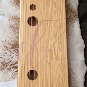 May include: A rectangular wooden lap harp with a light tan wood grain finish, featuring nineteen vertical metal strings, a dark brown bridge at the base, and four circular sound holes of varying sizes arranged along the left side of the instrument.