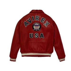 Op de afbeelding: Een rode leren jas met het woord "AVIREX" over de bovenrug, boven een cirkelvormig embleem en het woord "USA". De jas heeft een kraag, geribde manchetten en een geribde zoom met witte strepen.