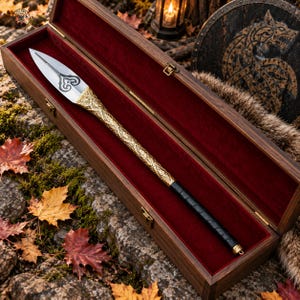 May include: A metal spearhead with a silver blade featuring a dark decorative engraving, a gold-toned shaft with intricate etched patterns, and a black leather-wrapped handle, presented inside a rectangular wooden box lined with red velvet.
