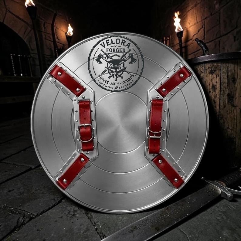 May include: A circular silver metal shield featuring two red leather straps with metal buckles and rivets. The center displays a black circular logo with the text VELORA FORGED KNIVES AXES SWORDS HANDCRAFTED ARTISANRY and an anvil with crossed axes.