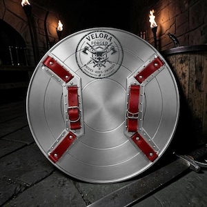 May include: A circular silver metal shield featuring two red leather straps with metal buckles and rivets. The center displays a black circular logo with the text VELORA FORGED KNIVES AXES SWORDS HANDCRAFTED ARTISANRY and an anvil with crossed axes.