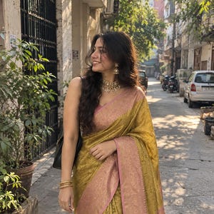 May include: A mustard yellow saree featuring a textured floral pattern and a wide, pale pink border with intricate gold zari work. The garment is paired with a matching pale pink sleeveless blouse, gold bangles, and a gold choker necklace with earrings.