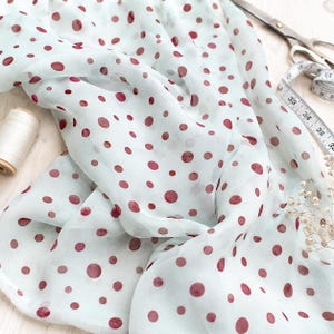 May include: A sheer, light mint green fabric featuring a pattern of dark red polka dots. The fabric is draped near a spool of white thread, a pair of metal scissors, and a white measuring tape showing measurements in inches.