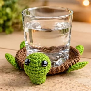 Puede incluir: Un vaso de agua transparente sobre un posavasos redondo de ganchillo con forma de tortuga. El posavasos tiene un centro marrón, una cabeza verde brillante con grandes ojos de plástico negros y blancos, y cuatro pequeñas aletas verdes.