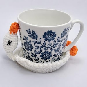 Peut inclure: Une tasse en céramique blanche avec un motif floral bleu est posée sur un dessous de verre crocheté blanc. Le dessous de verre présente un motif d'oie crochetée, avec un bec et des pieds orange, et une croix noire pour un œil.