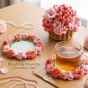 May include: A Blushing Blooms Coaster Set featuring a crochet coaster with a floral design in shades of pink, coral, and green. A matching crochet flower basket and a cup of tea are also present. The set is handmade and adds a touch of elegance.