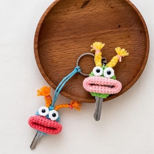 Könnte beinhalten: Zwei handgefertigte Häkel-Schlüsselanhänger, jeweils mit einem skurrilen Monster-Design. Einer hat einen blauen Kopf mit orangefarbenen Akzenten, der andere einen grünen Kopf mit gelben Akzenten. Beide haben große Kulleraugen und rosa Münder.
