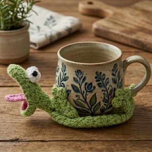 Könnte beinhalten: Eine Keramiktasse mit floralem Muster wird von einem grünen gehäkelten Alligator umschlungen. Der Alligator hat ein weißes Auge und eine rosa Zunge. Die Tasse steht auf einer Holzoberfläche, mit einer kleinen Topfpflanze und einem gemusterten Tuch im Hintergrund.