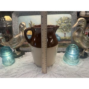 May include: A brown and beige ceramic pitcher with a handle, measuring approximately 9 inches tall. The pitcher is displayed with decorative items, including glass insulators and metal duck figurines. A ruler is placed next to the pitcher.