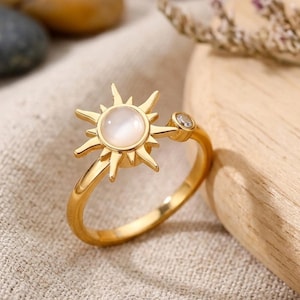 May include: A gold ring with a sunburst design and a central, round, pearlescent stone. The ring has a delicate band and a small, clear gemstone accent. The ring is set against a neutral background with decorative elements.