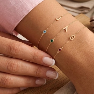 Puede incluir: Tres delicadas pulseras de cadena dorada, cada una con un pequeño dije de letra y una diminuta piedra preciosa redonda. Las pulseras muestran las letras J, A y S, combinadas con piedras de color azul claro, verde y rojo respectivamente.