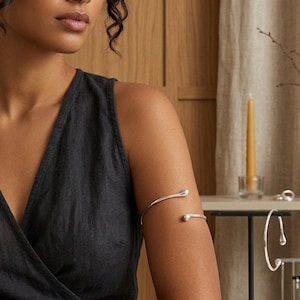 May include: A polished silver arm cuff featuring a minimalist open design with rounded, teardrop-shaped ends. The sleek metal band curves smoothly around the upper arm, showcasing a reflective surface and a simple, continuous metallic structure.