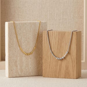 May include: Two necklaces on display. One is gold-coloured, featuring a bar and bead design, the other is silver-coloured with a similar bar and bead design. Both have fine chains and are shown against a neutral backdrop.