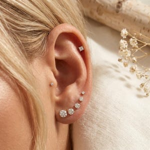 May include: A set of seven gold-toned stud earrings featuring clear, round-cut stones of varying sizes. One square-cut stud is positioned in the upper cartilage, while the remaining six round studs are arranged in a line along the earlobe.