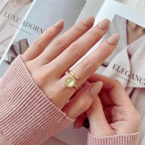 May include: A gold-colored ring with a round, light-colored stone and a small diamond accent. The ring is on a finger, and the design includes a spiral band. The ring is a piece of jewelry.
