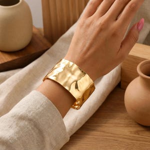 Può includere: Un ampio bracciale a fascia color oro caratterizzato da una superficie martellata, irregolare e altamente riflettente. Il cinturino in metallo presenta un bordo curvo e liscio con una finitura lucida che cattura la luce, creando profondi riflessi.