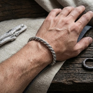 May include: A silver-toned metal cuff bracelet featuring a twisted rope design. The bracelet has a polished finish with darkened recessed areas between the twisted strands, creating a textured appearance around the entire circumference of the band.