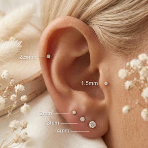 May include: A set of five round, clear stud earrings worn in various ear piercings. The earrings are labeled with sizes: 2.5mm for the helix, 1.5mm for the tragus, and 2mm, 3mm, and 4mm for the lobe piercings, showing the scale of each stone.