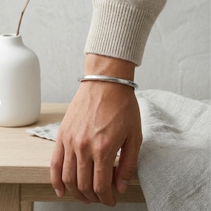 May include: A slim, polished silver-tone metal cuff bracelet with a smooth, rounded exterior surface. The bracelet features subtle, small engraved lettering along the outer band, creating a clean and understated look on the wrist.