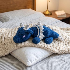 May include: A handmade blue crochet dragon plush toy with gray accents, lying on a cream-colored blanket over a white pillow. The dragon has horns and a friendly expression. The background includes a bed with striped sheets and a wooden nightstand.