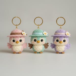 May include: Three crochet bird keychains in pastel colors: pink, mint green, and lavender. Each bird wears a matching hat with a flower accent and has a gold-colored keyring. The birds have large eyes and small wings.