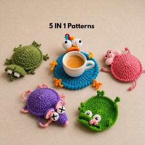 May include: A collection of five handmade crochet coasters in various whimsical creature designs. The coasters include a green frog, a pink pig, a purple mouse, a green monster, and a blue coaster with a bird design holding a white coffee cup. The text "5 IN 1 Patterns" is visible.