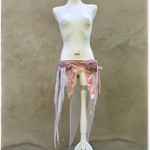 Puede incluir: Una falda de disfraz rosa y blanca con detalles florales y tela fluida, exhibida en un maniquí blanco. La falda presenta un material similar al satén y detalles delicados y en capas, creando un aspecto caprichoso y etéreo.